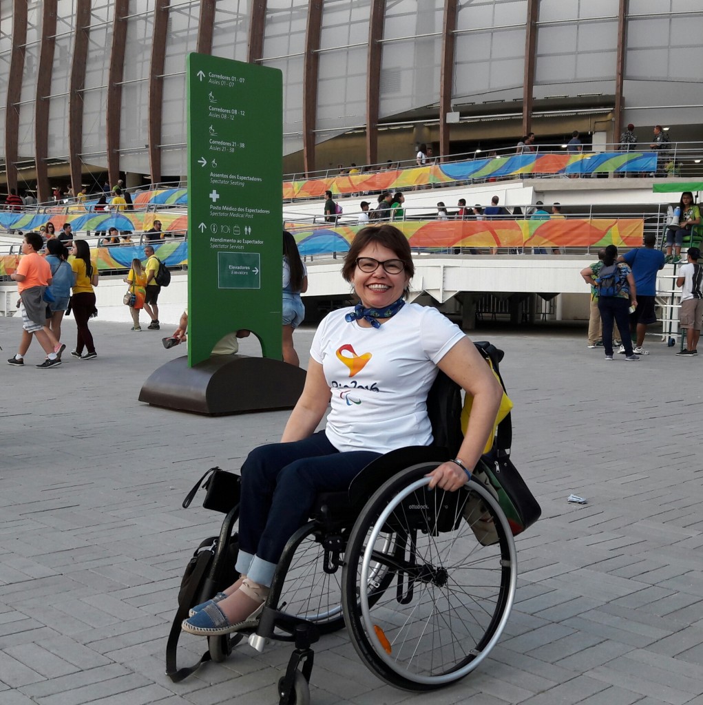 Estou em frente à Arena Olímpica 3, para assistir ao jogo de bocha. Estou vestida com a camisa que contém a logo da Paralimpíada e com bandana com as cores da bandeira brasileira.