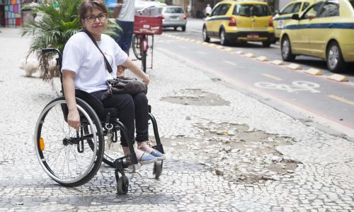 Laura aponta buracos em uma calçada portuguesa. Ela está em sua cadeira de rodas.