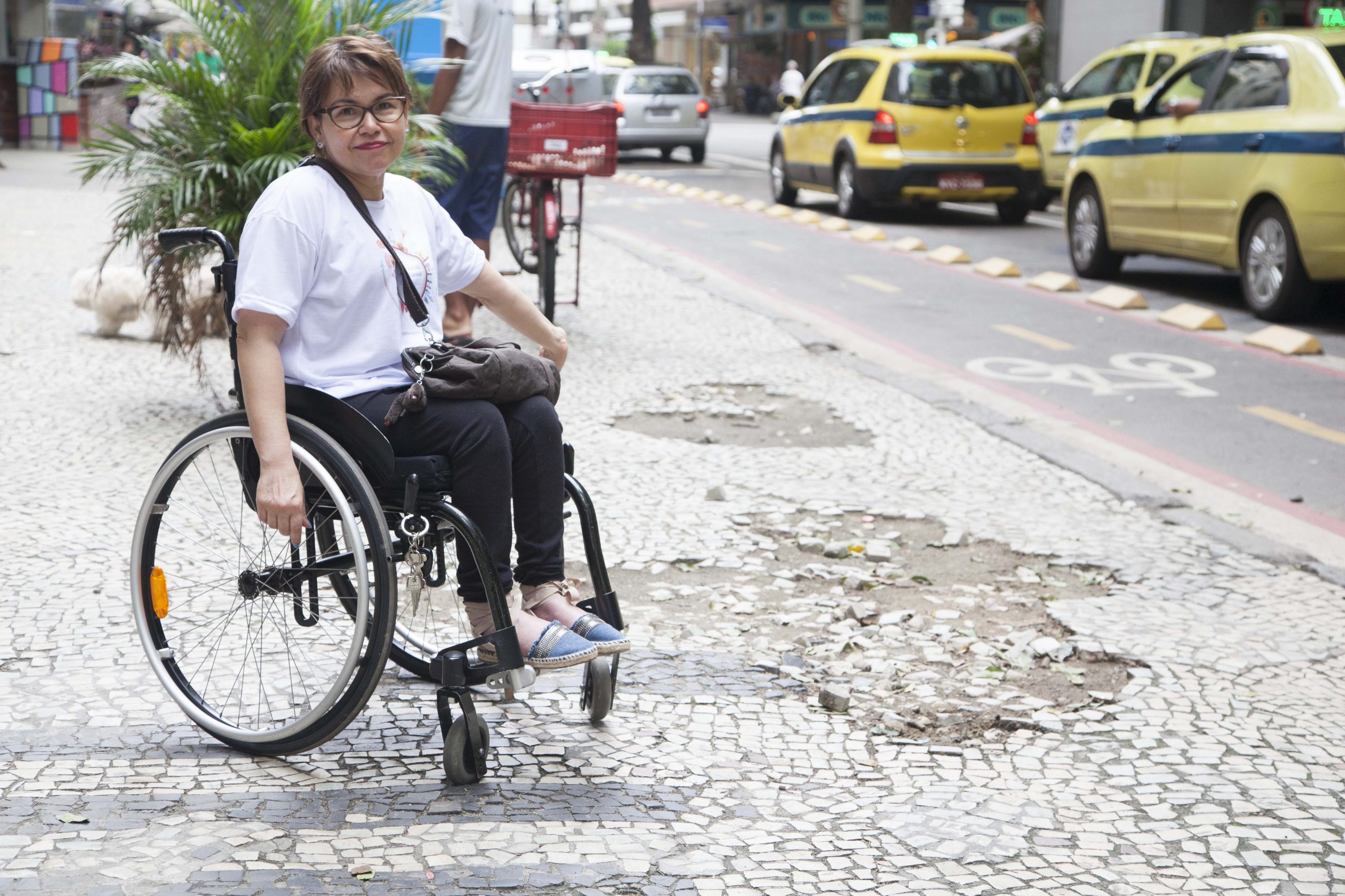 Laura aponta buracos em uma calçada portuguesa. Ela está em sua cadeira de rodas.