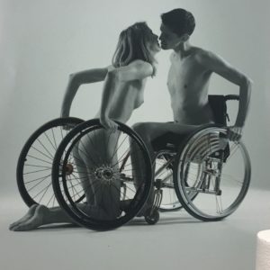 Um homem e uma mulher se beijam. Ele está à direita, assentado na cadeira de rodas. Ela está ajoelhada à esquerda, segurando duas rodas de cadeira, sendo uma de cada lado.