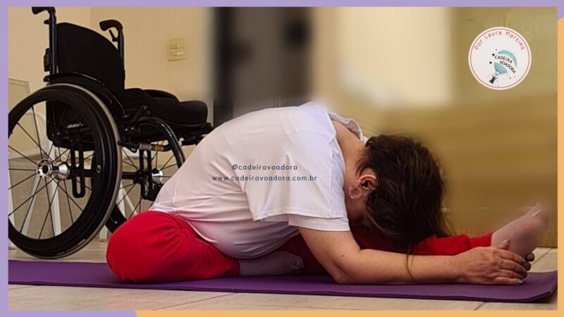 Laura está assentada no chão, realizando uma postura de yoga. Atrás dela, está sua cadeira de rodas.