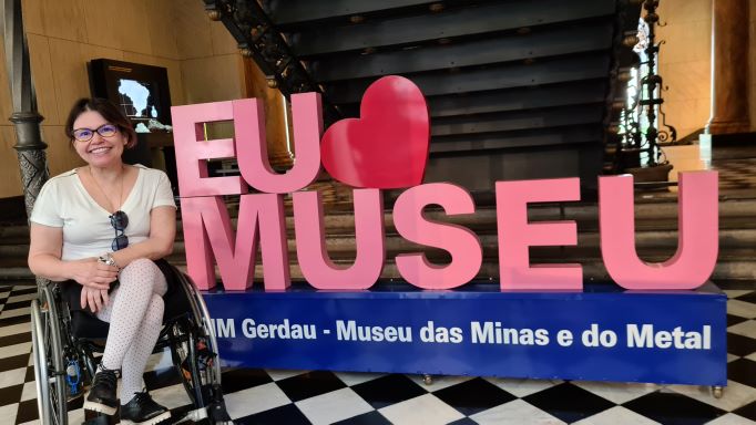 Laura está em frente a uma instalação em que está escrito "Eu amo (c/ um coração) Museu das Minas e do Metal