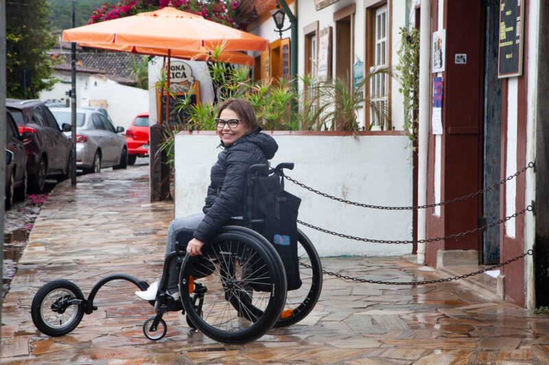 Laura está sob chuva fina. Sua cadeira de rodas tem uma 5a roda acoplada na frente, entre os pedais. Esta roda eleva as rodinhas dianteiras, tornando a circulação mais fácil.