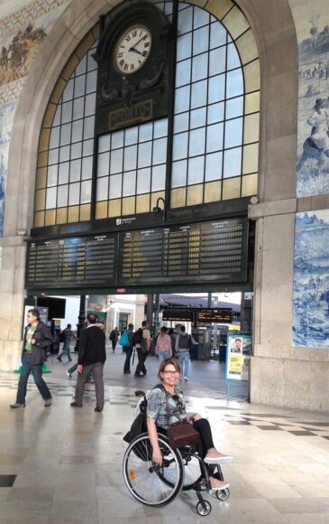Laura posa pra foto em frente ao pórtico de entrada para a área onde partem os trens. No alto, há um relógio analógico. Ao fundo, pessoas circulando.