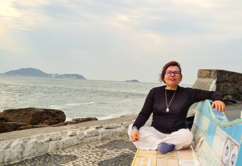 Laura está assentada em um banco revestido com azulejos coloridos. Ela está com as pernas cruzadas em posição de yoga e tem , ao lado, parte do calçadão de pedra portuguesa e, ao fundo, o mar, com montanhas em segundo plano.